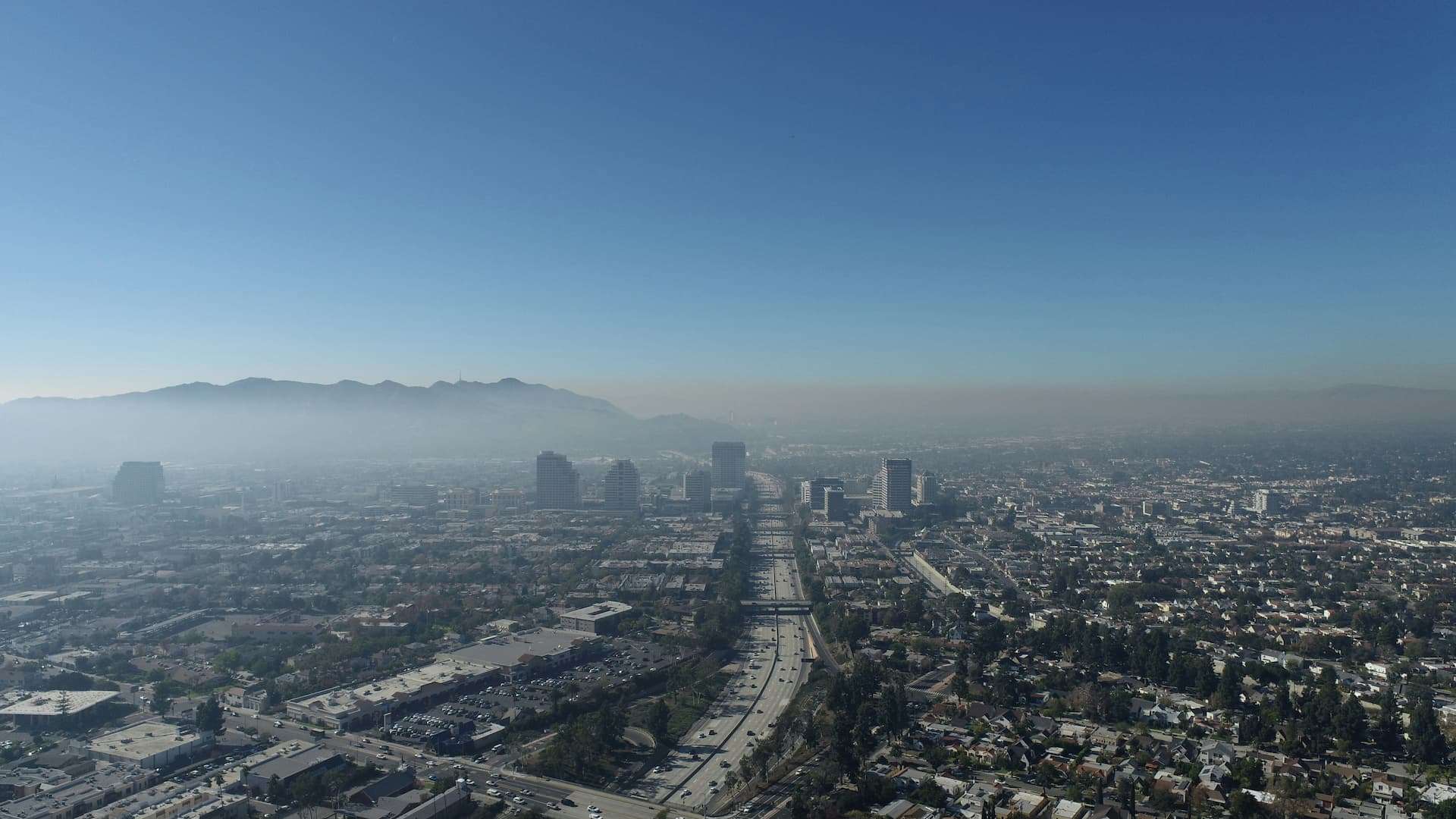 Wide cityscape with a clear blue sky, tall buildings near a highway, and distant mountains on the horizon.